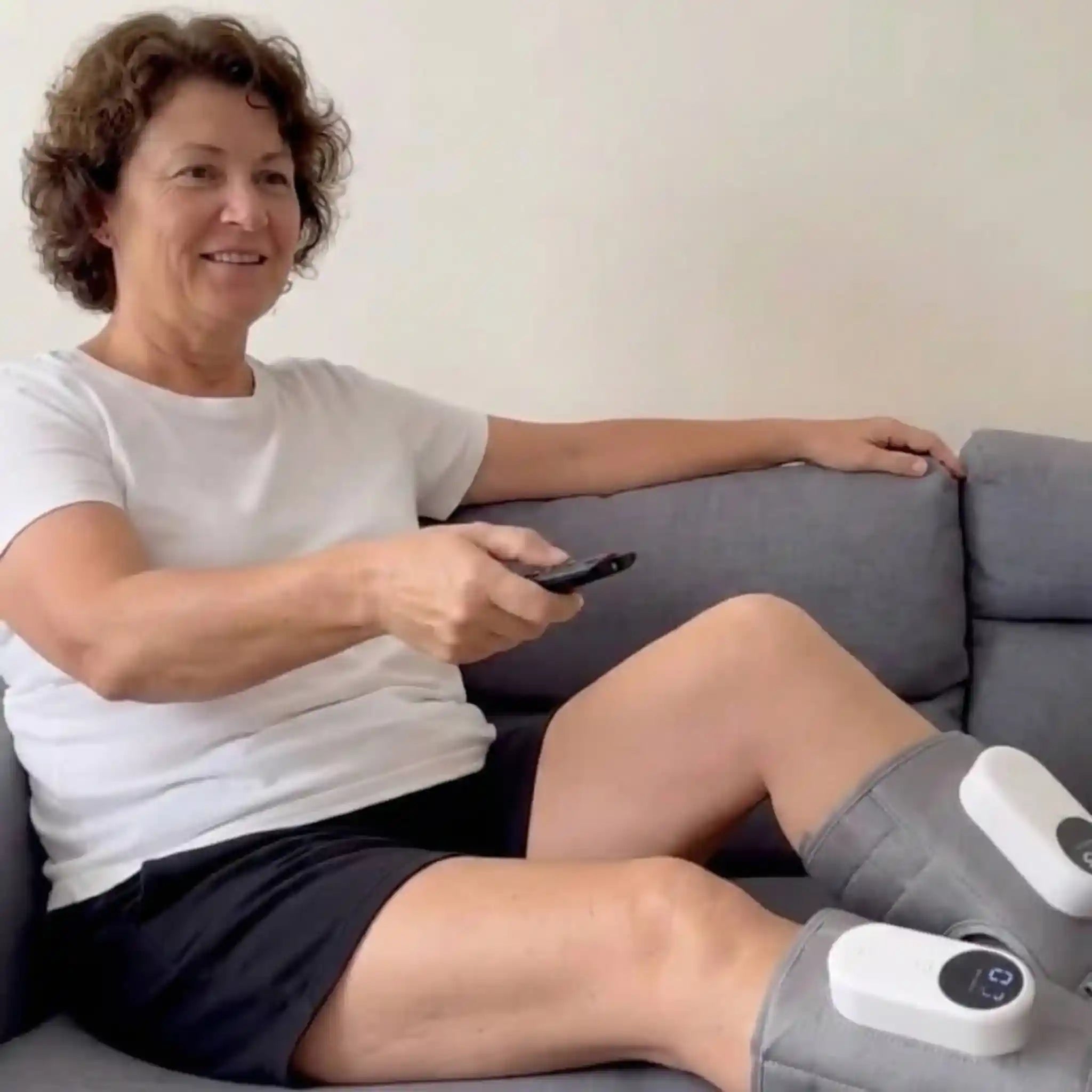 Woman holding TV remote and smiling while wearing Recovie leg massager on couch